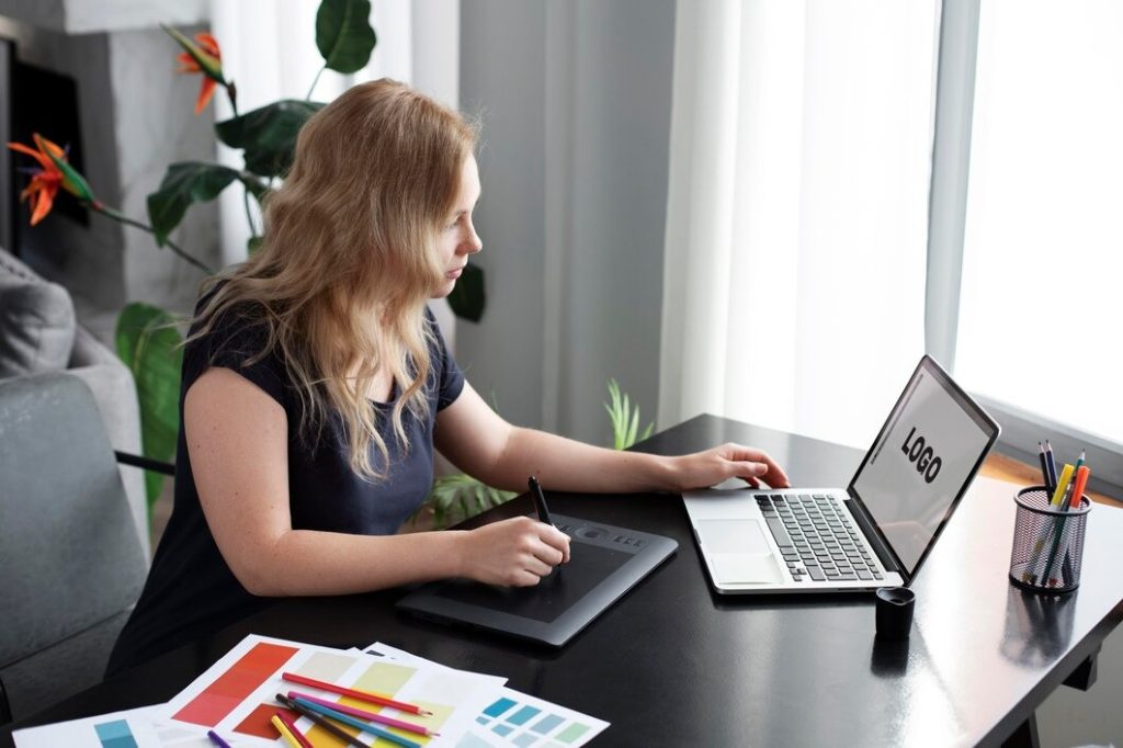 A woman sits at a table with a laptop open and a pen in her hand, focused on her work
