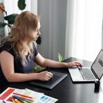 A woman sits at a table with a laptop open and a pen in her hand, focused on her work