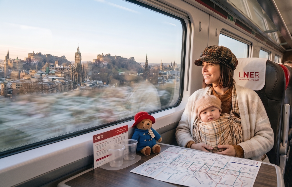 Woman and baby enjoy scenic train journey through Edinburgh, with plush bear and map on table in LNER carriage