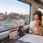 Woman and baby enjoy scenic train journey through Edinburgh, with plush bear and map on table in LNER carriage